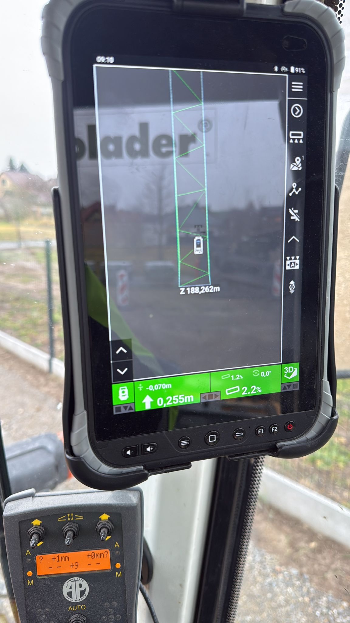 Ekran tabletu z mapą 3D i danymi pomiarowymi w kabinie maszyny budowlanej, prawdopodobnie podczas utwardzania drogi lub układania kostki. Widoczne panele sterowania.