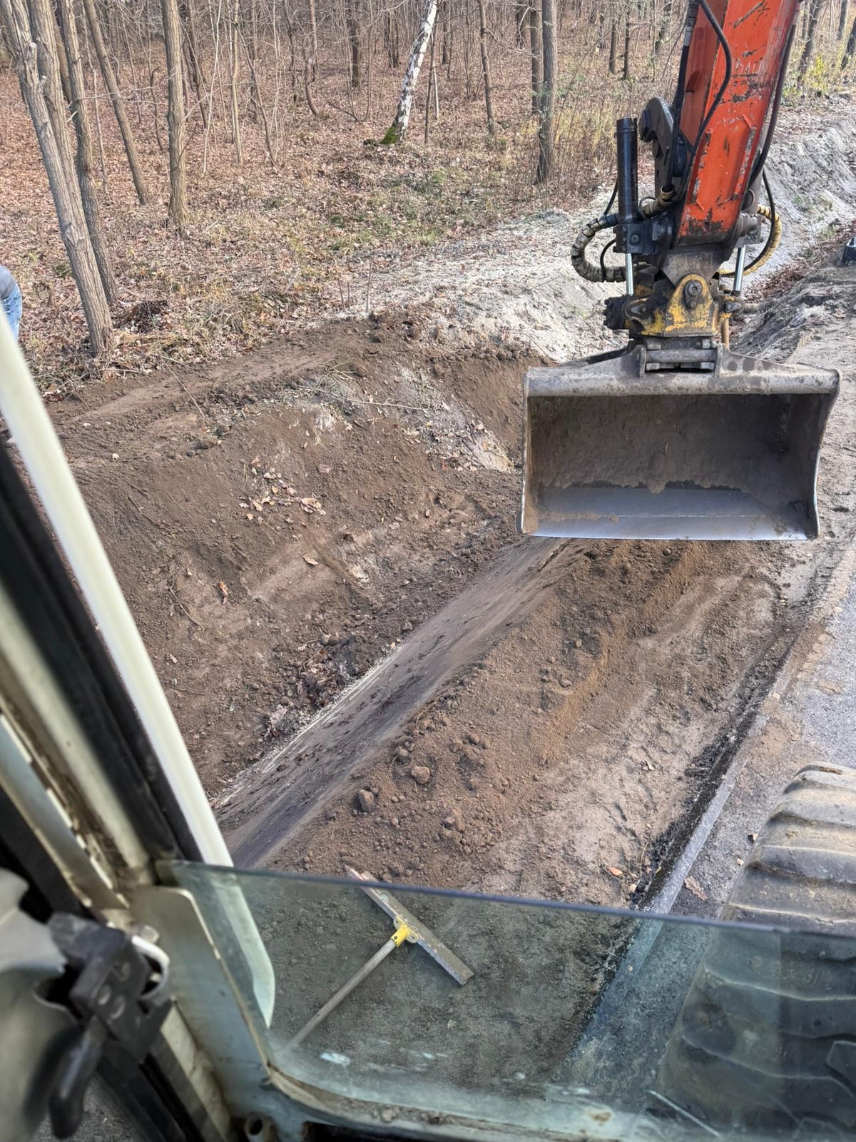 Koparka w trakcie prac ziemnych, widok z kabiny operatora. Wykopany rów w ziemi, w tle las. Przygotowanie terenu pod utwardzenie drogi.