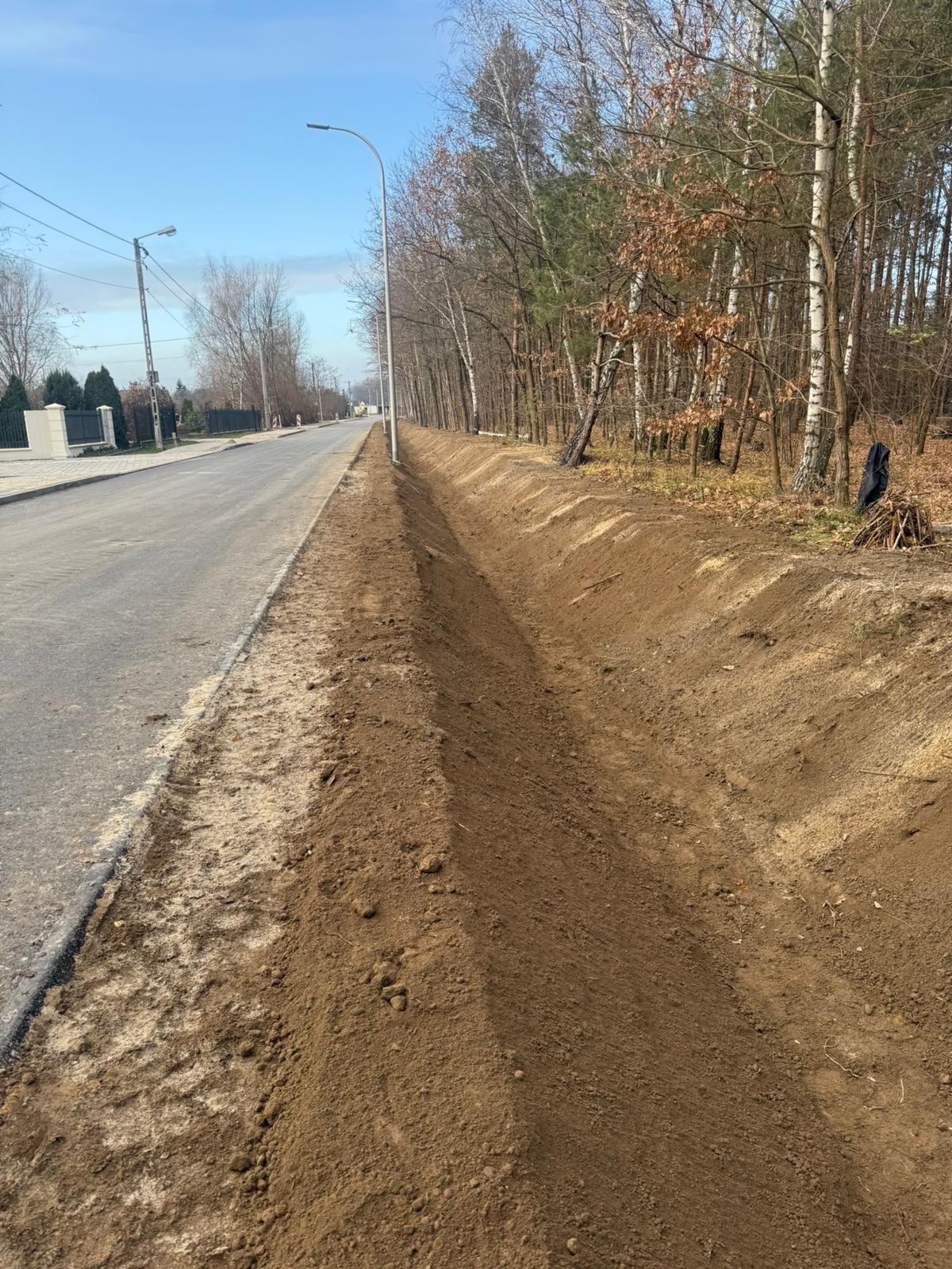 Przygotowany teren pod utwardzenie pobocza drogi, widoczny wykop wzdłuż asfaltu, drzewa w tle, słup oświetleniowy.