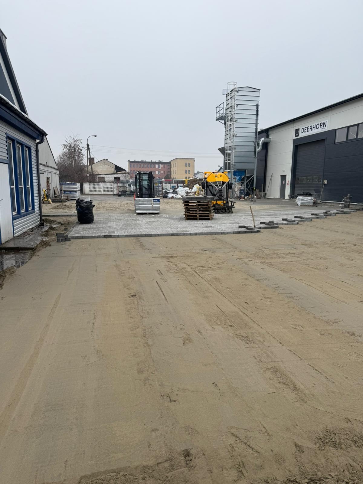 Prace brukarskie w toku: mini koparka, palety z kostką i fragment ułożonej nawierzchni na placu budowy. W tle budynki i elementy infrastruktury.