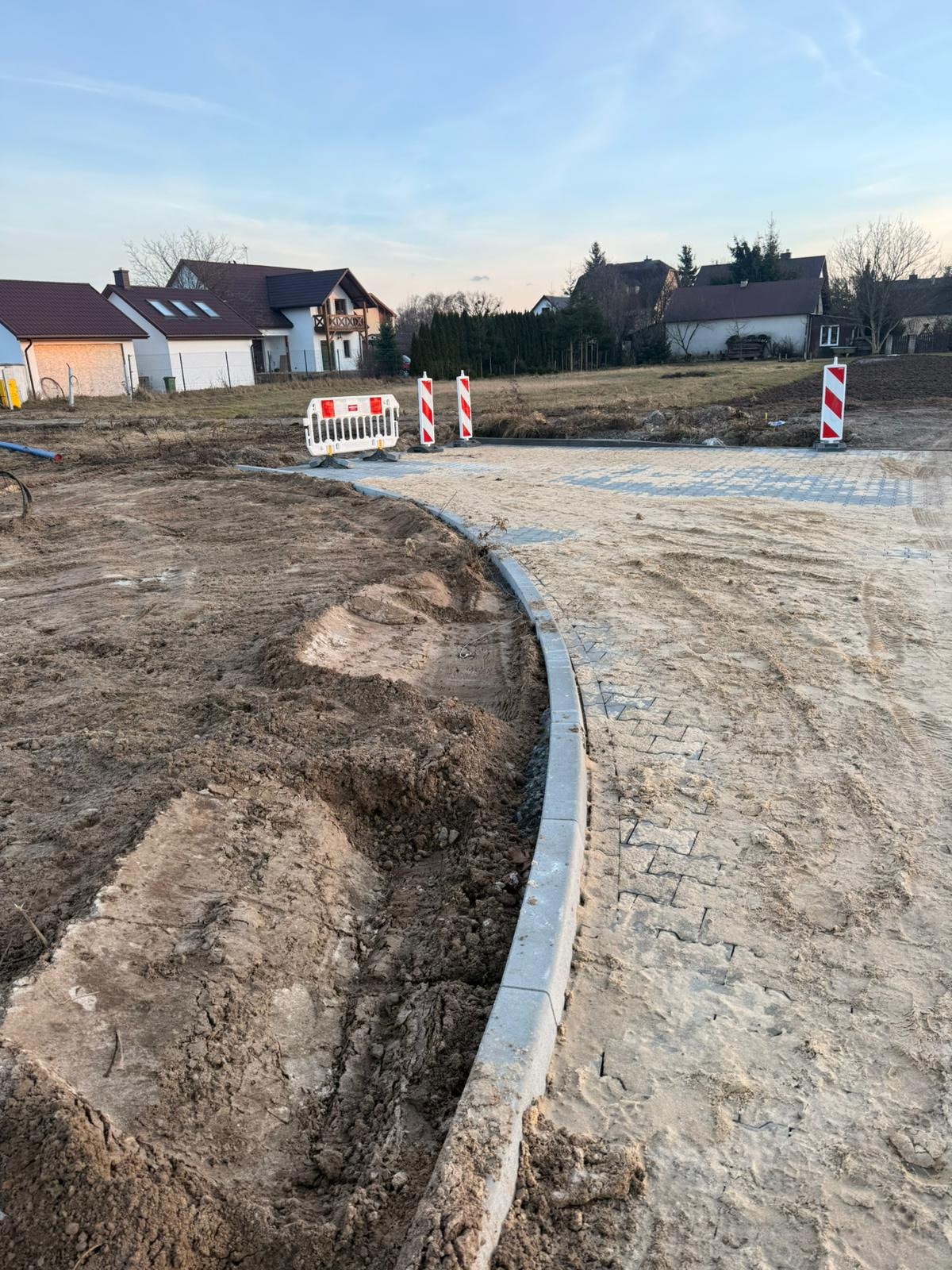 Etap układania kostki brukowej: ułożone obrzeża, częściowo wyłożona kostka, teren przygotowany pod dalsze prace. Widoczne zabezpieczenia budowy.