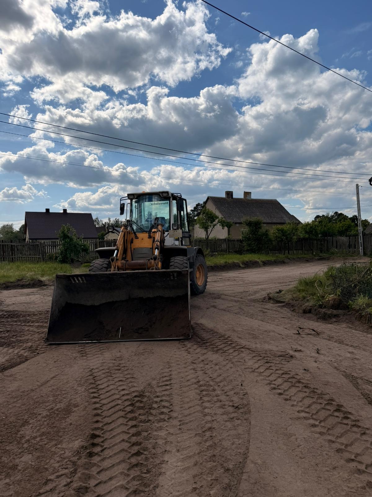 Żółta ładowarka na gruntowej drodze z widocznymi śladami opon, w tle domy i błękitne niebo z obłokami. Maszyna budowlana przygotowuje podłoże pod budowę.