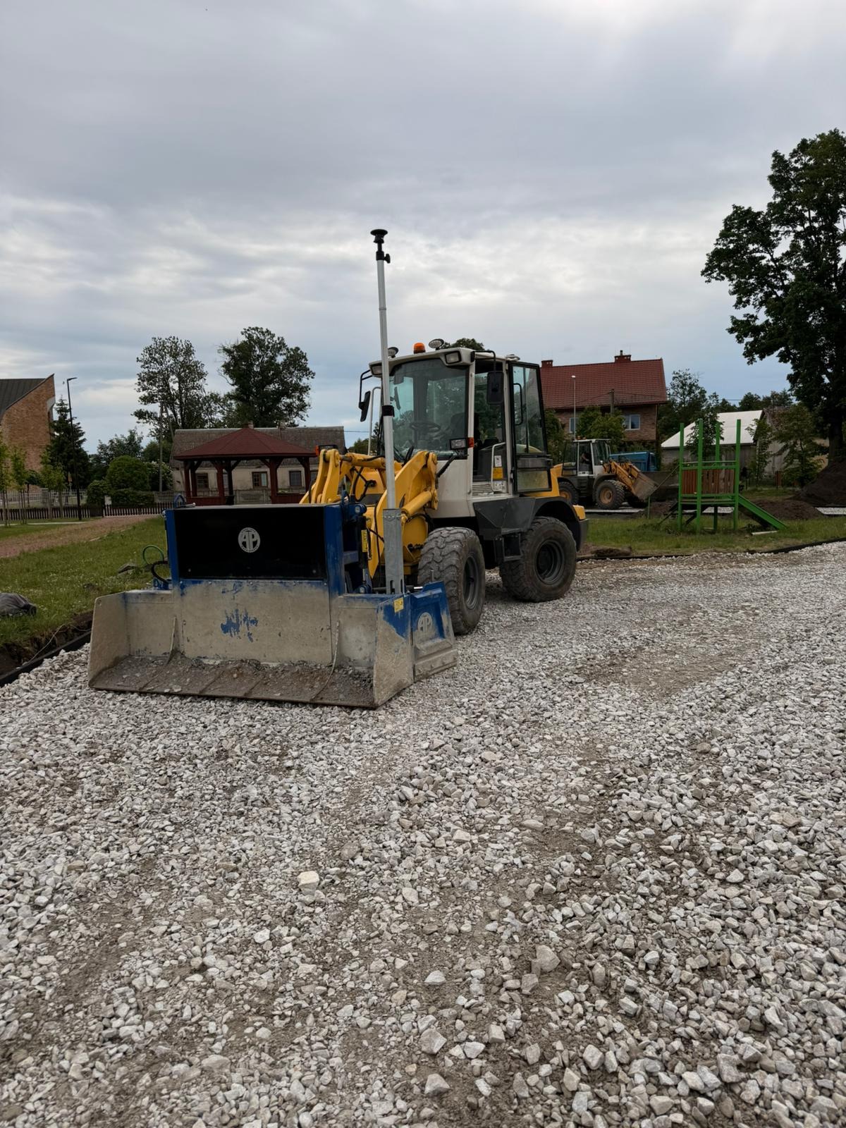 Żółto-szara równiarka z czarnym panelem na żwirowej nawierzchni. W tle budynki i zieleń, pochmurne niebo. Etap prac przygotowawczych podłoża.