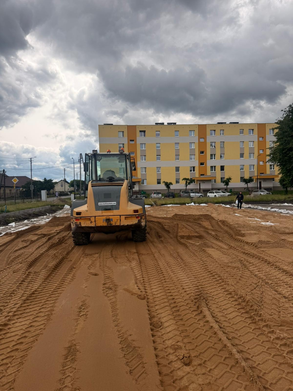 Żółty walec drogowy ubija piasek na placu budowy, w tle blok mieszkalny i pochmurne niebo. Widoczne ślady opon na piasku.