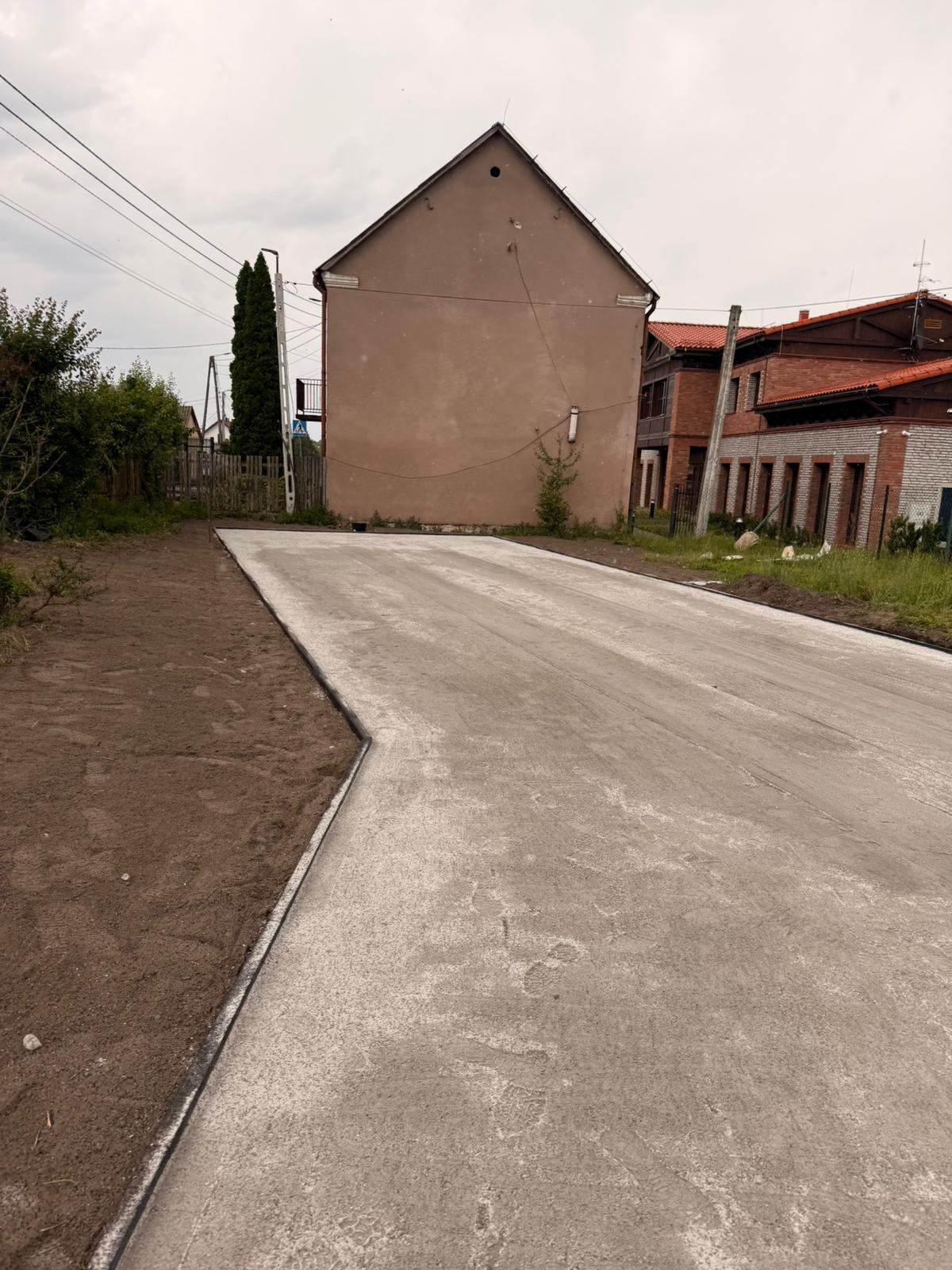 Nowa, betonowa nawierzchnia drogi z obrzeżami, świeżo wylana, z widoczną fakturą betonu. W tle budynki mieszkalne i zieleń. Po lewej stronie widoczna ziemia.