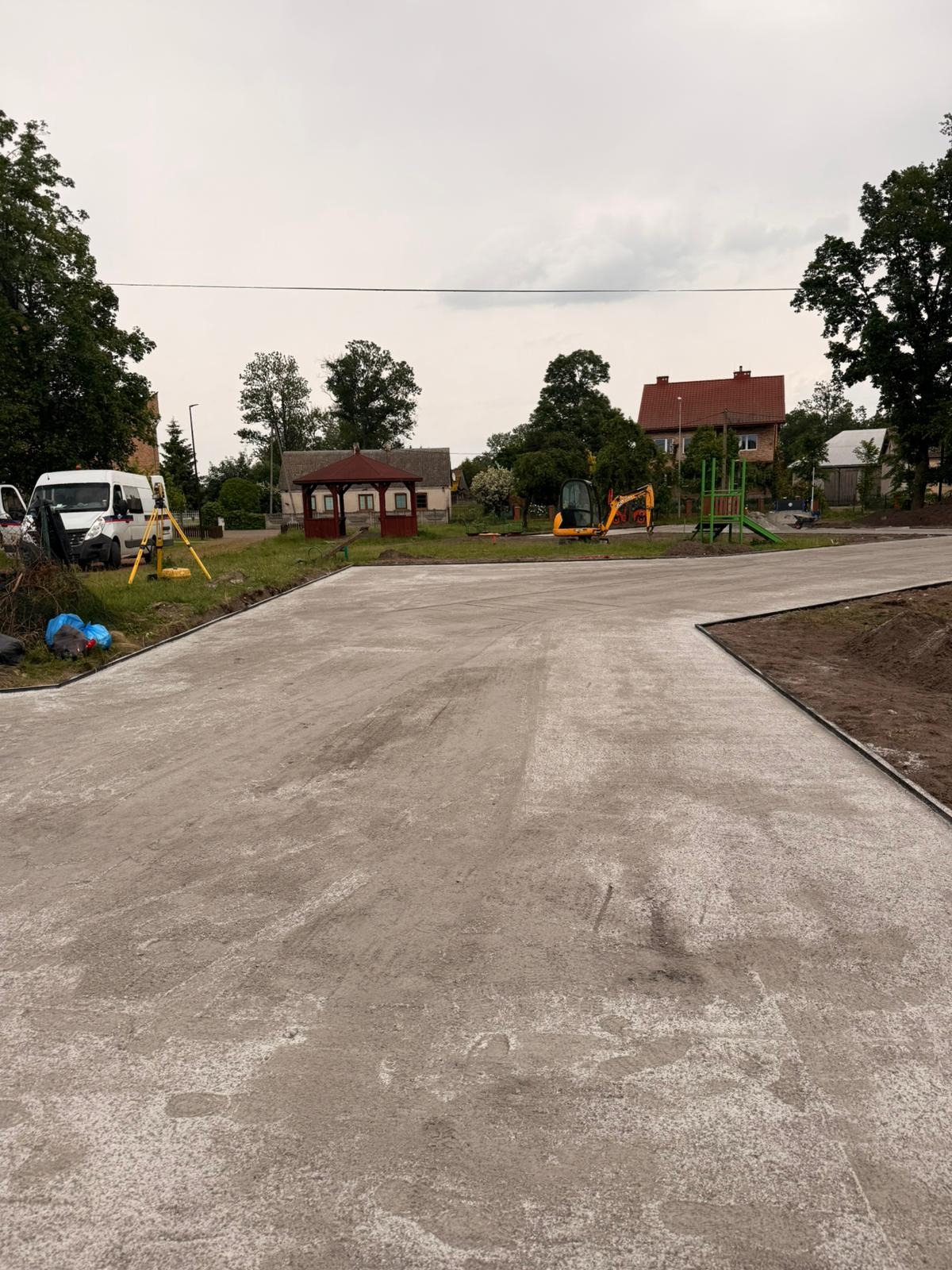Wykończona, utwardzona nawierzchnia w Sól, widok z poziomu gruntu. W tle zabudowa wiejska, mini koparka i niwelator. Szary dzień.