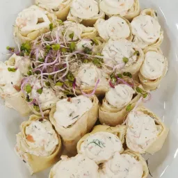 Roladki z tortilli wypełnione kremowym farszem z ziołami i łososiem, udekorowane świeżymi kiełkami rzodkiewki, ułożone na białym talerzu.