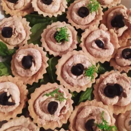 Tarty z kremowym nadzieniem i czarną oliwką, ułożone na zielonych liściach, widoczne z góry.