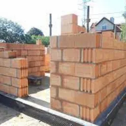 Wznoszenie ściany z czerwonych, porowatych bloczków ceramicznych. Widoczne czarne zabezpieczenie fundamentu i fragment budynku w tle.