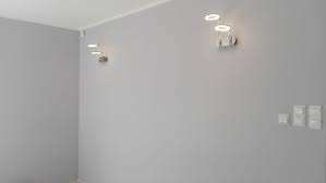 Szare, gładkie ściany w rogu pokoju z trzema okrągłymi lampami ściennymi i białymi włącznikami światła. Widoczny biały sufit z listwą przypodłogową.