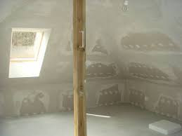 Poddasze po wstępnym etapie prac wykończeniowych, widoczne nierówności i pozostałości po szpachlowaniu na ścianach skośnych, drewniana belka pionowa na pierwszym planie, małe okno dachowe w tle.