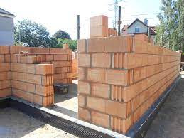 Wznoszenie ściany z czerwonych, porowatych bloczków ceramicznych. Widoczne czarne zabezpieczenie fundamentu i fragment budynku w tle.