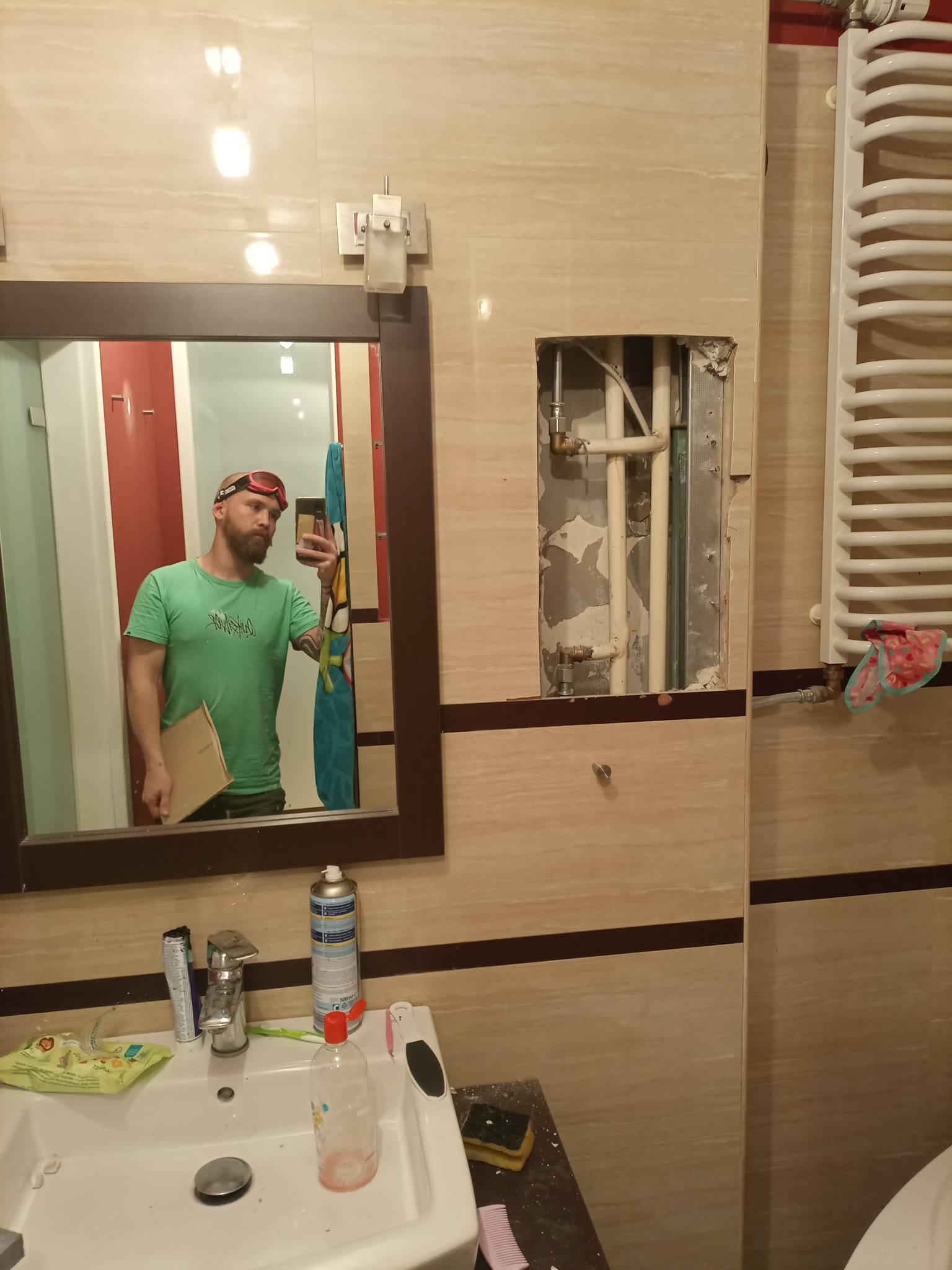 Remont łazienki w trakcie: widoczne rury instalacji grzewczej w ścianie, grzejnik drabinkowy, oraz odbicie w lustrze osoby robiącej zdjęcie, ubrana w okulary ochronne i trzymająca kartkę.