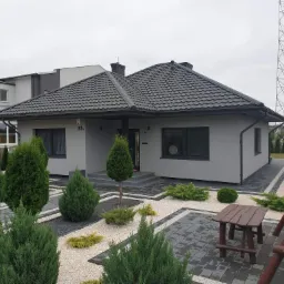 Szary, parterowy dom z ciemną dachówką i numerem 83a, otoczony ozdobnym ogrodem z jasnym żwirem, ciemnymi obrzeżami z kostki brukowej i iglakami.