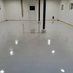Świeżo wylana, szara posadzka żywiczna w jasnym pomieszczeniu z dwoma filarami, widoczne małe, łukowe okna i białe ściany w stanie surowym.