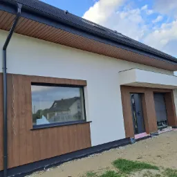 Nowoczesny dom jednorodzinny z elewacją w odcieniach bieli i drewna. Widoczna stolarka okienna w kolorze czarnym oraz fragment dachu z czarną dachówką.