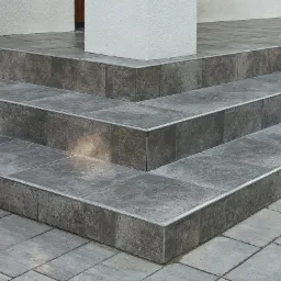 Schody wejściowe wyłożone szarą, kwadratową płytką z metalowymi listwami krawędziowymi, prowadzące do budynku z jasną elewacją i kolumną.