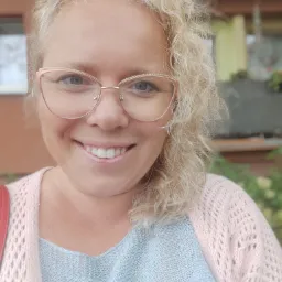 Portret uśmiechniętej kobiety z blond kręconymi włosami i okularami w różowych oprawkach, prezentującej białe, równe zęby.