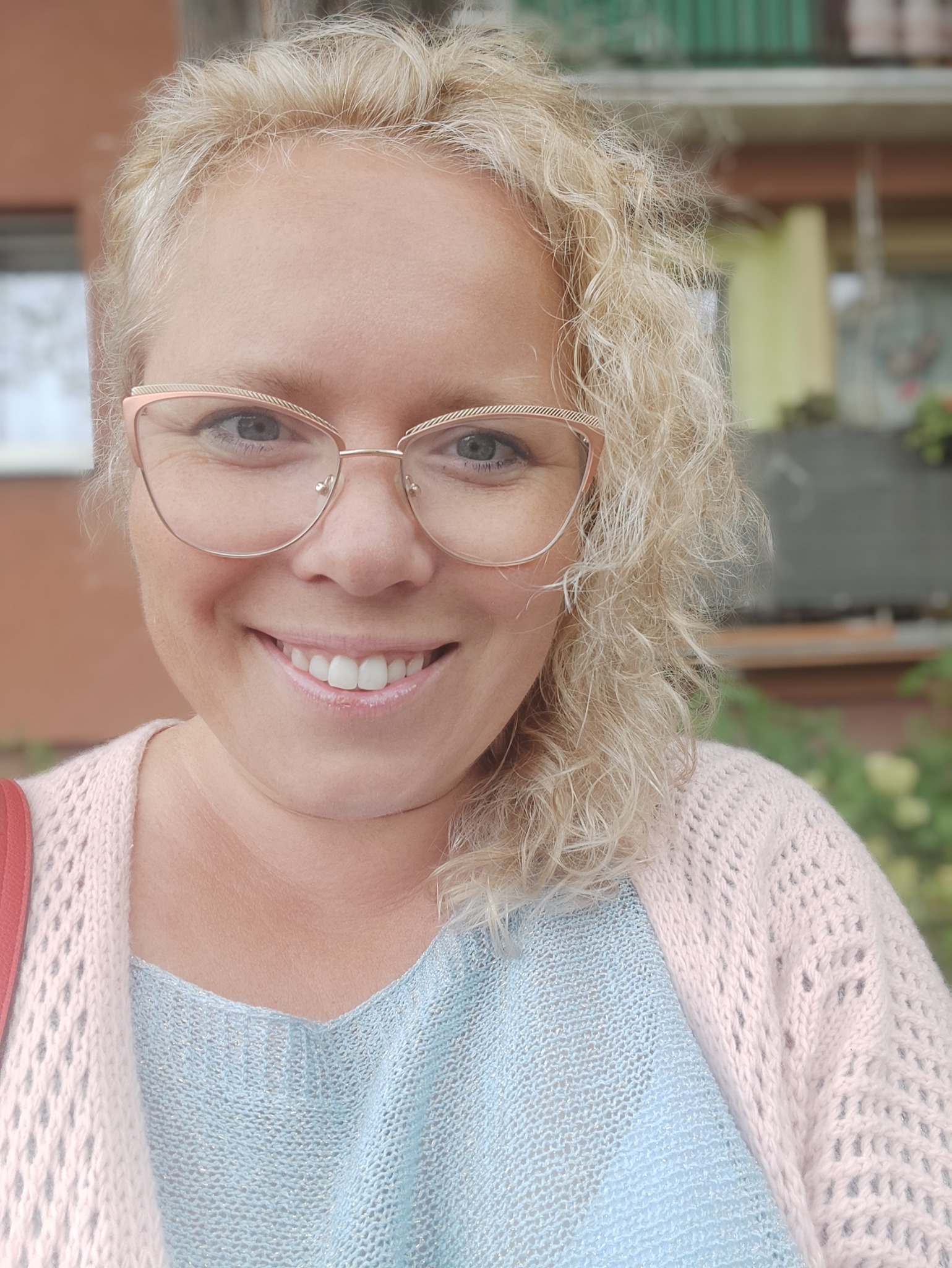 Portret uśmiechniętej kobiety z blond kręconymi włosami i okularami w różowych oprawkach, prezentującej białe, równe zęby.