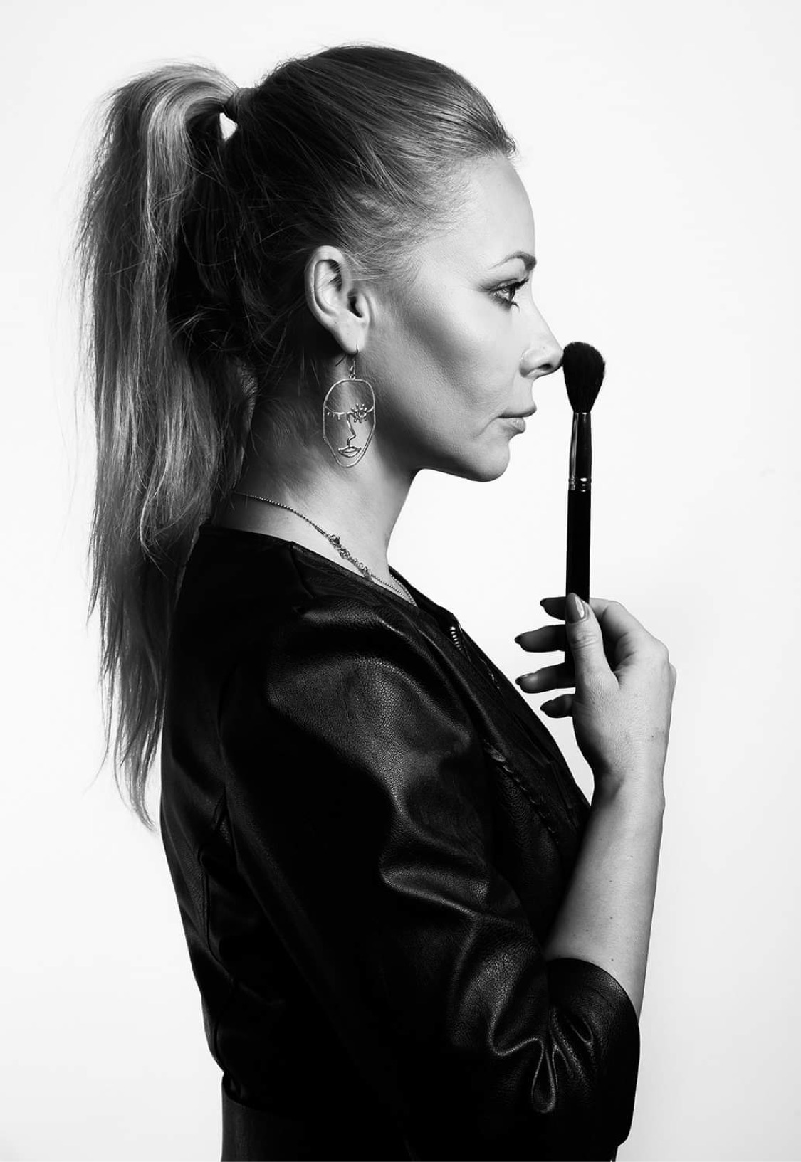 Portret kobiety z pędzlem do pudru dotykającym nosa, profil, kolczyk z motywem twarzy, czarno-białe zdjęcie.