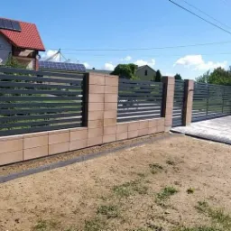 Nowoczesne ogrodzenie posesji w Szczuczynie z przesuwną bramą, wykonane z poziomych, grafitowych profili i elementów z beżowego kamienia.