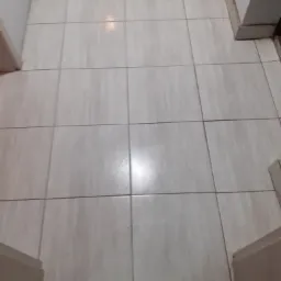 Beżowe, kwadratowe płytki podłogowe ułożone w regularną siatkę, widoczne fragmenty białych ścian i futryn drzwi.