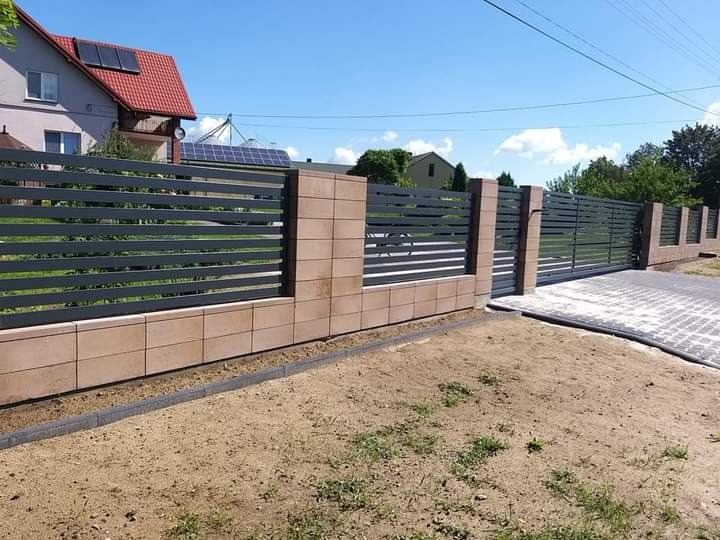 Nowoczesne ogrodzenie posesji w Szczuczynie z przesuwną bramą, wykonane z poziomych, grafitowych profili i elementów z beżowego kamienia.