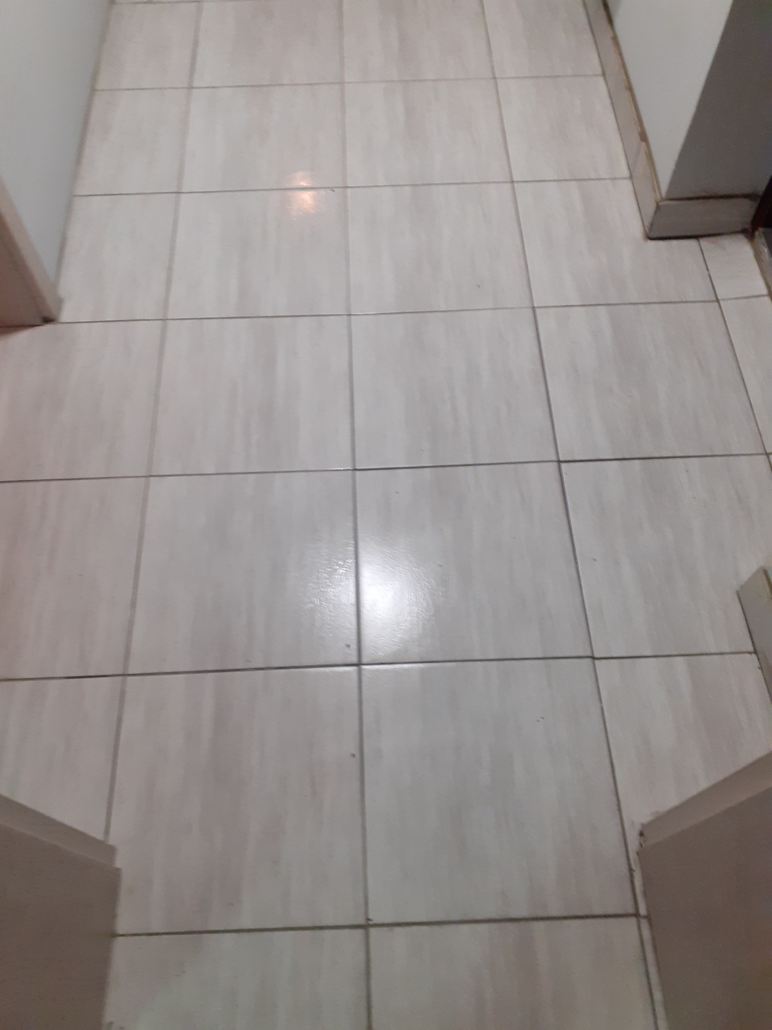 Beżowe, kwadratowe płytki podłogowe ułożone w regularną siatkę, widoczne fragmenty białych ścian i futryn drzwi.