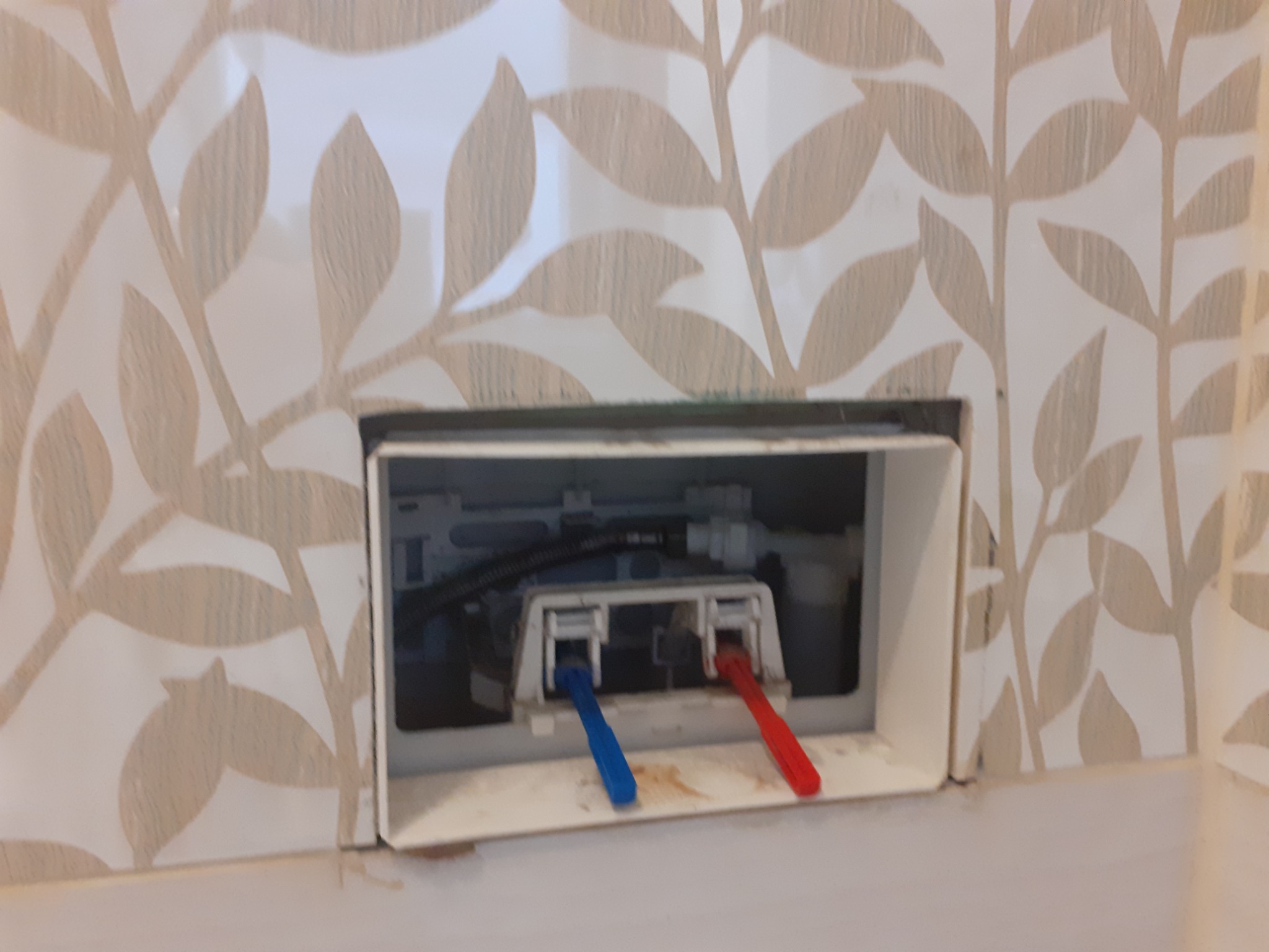 Podłączenie instalacji wodnej do spłuczki podtynkowej, widoczne wężyki zasilające wodą zimną i ciepłą, tło stanowi ściana wykończona płytkami ceramicznymi z motywem liści.