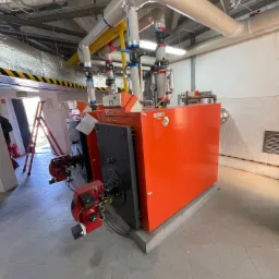 Pomarańczowy kocioł gazowy Viessmann z czarnymi elementami, podłączony do rur na tle szarej podłogi i ściany z widoczną instalacją sufitową.