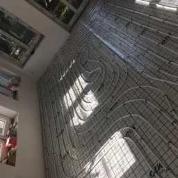 Białe rury ogrzewania podłogowego ułożone spiralnie na izolacji z nadrukowaną siatką, widoczne okna z roślinnością na zewnątrz, wpadające promienie słoneczne.