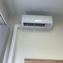 Biały klimatyzator Rotenso zamontowany na jasnej ścianie, widoczne rury instalacyjne i fragment żaluzji.