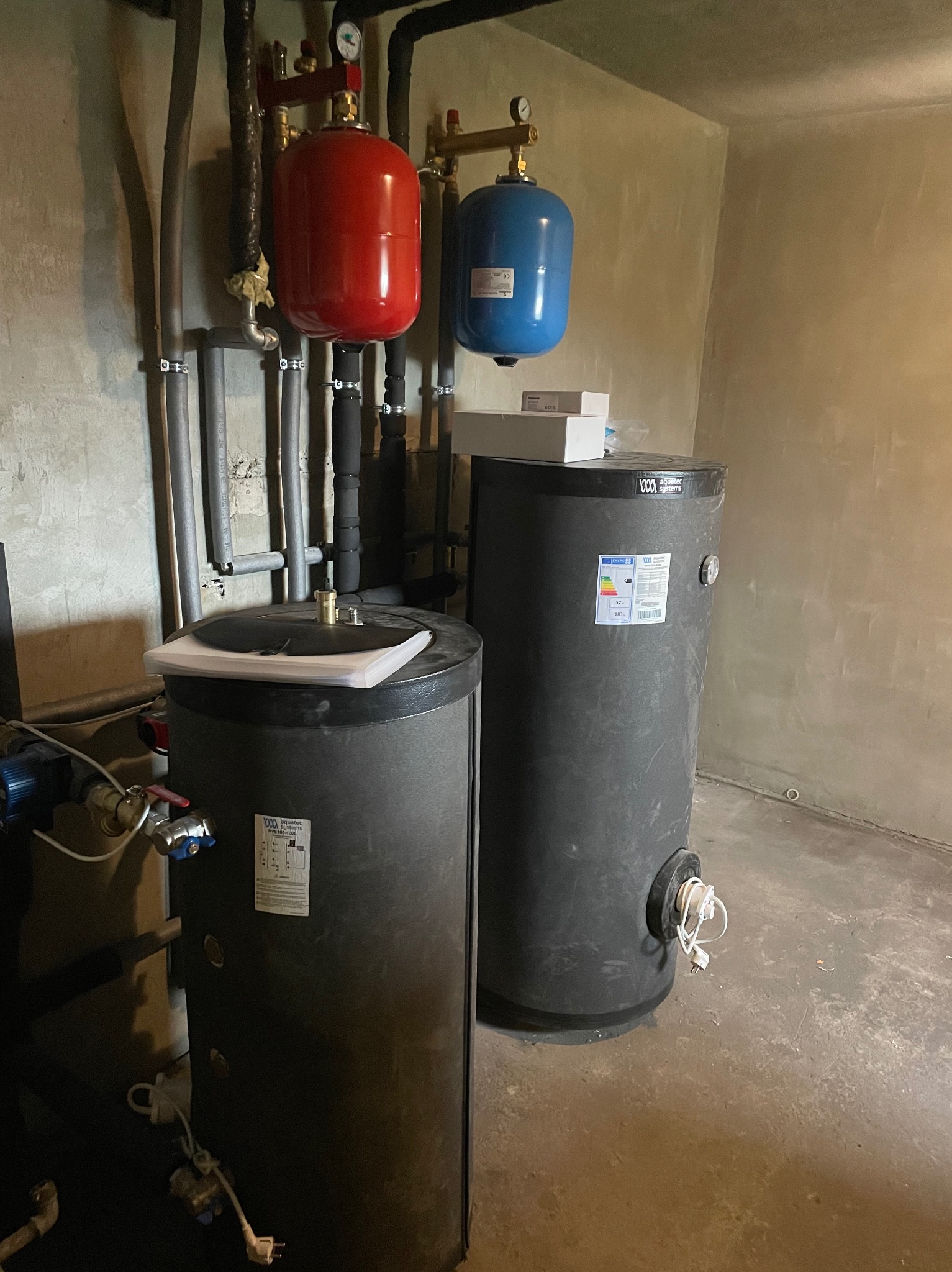 Dwa zbiorniki akumulacyjne ciepła (czerwony i niebieski) oraz dwa bojlery w kotłowni. Widoczne rury, zawory i manometry. Szare ściany i betonowa podłoga.
