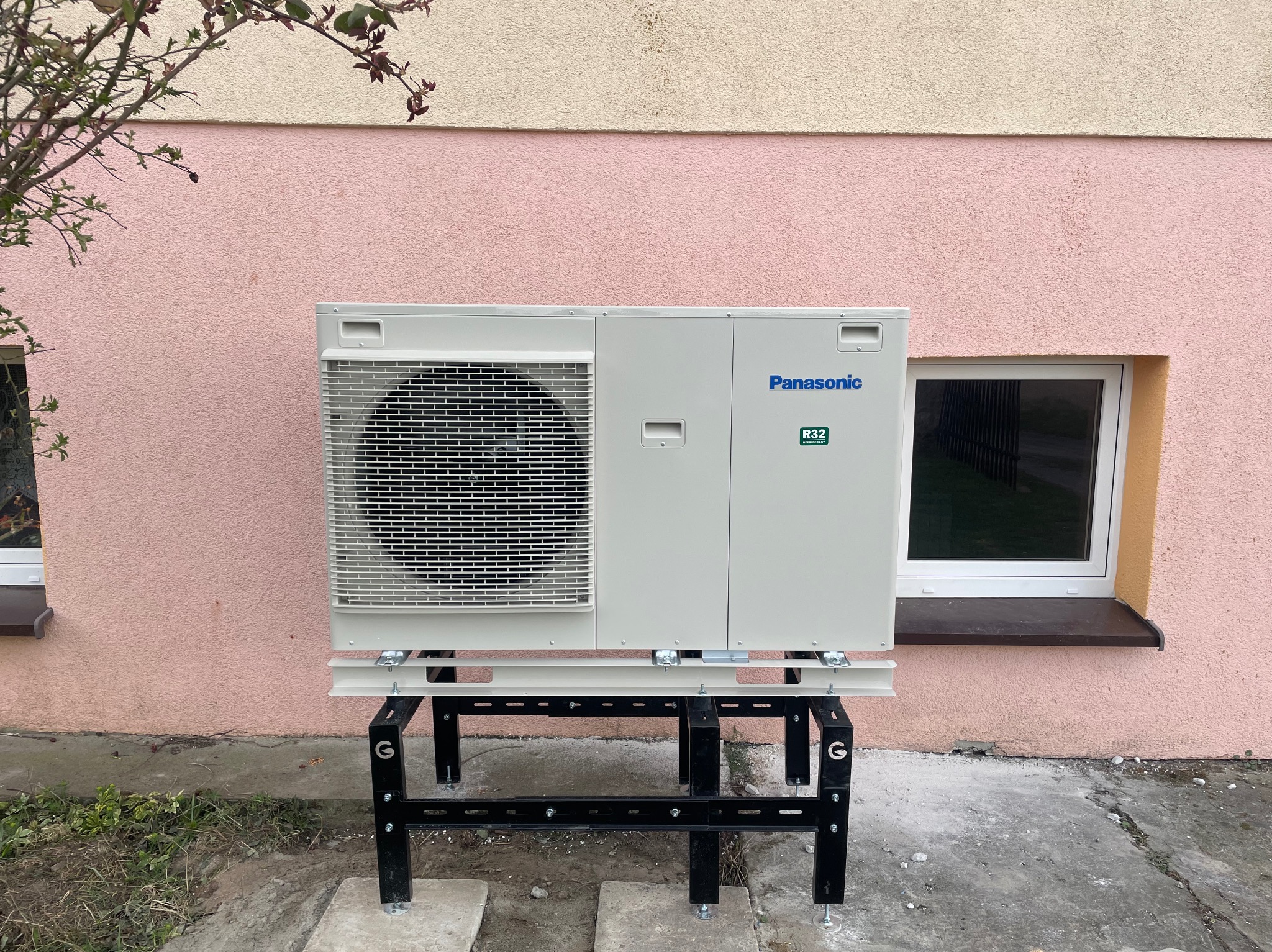 Biała pompa ciepła Panasonic na czarnym stelażu, zamontowana przy różowej ścianie budynku z oknem. Widoczny fragment drzewa.