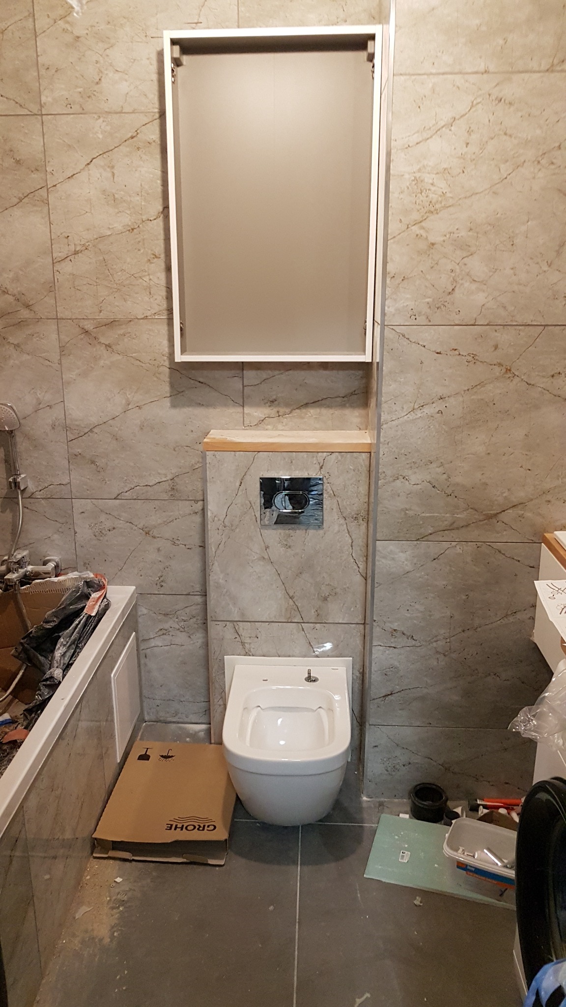 Wnętrze łazienki w trakcie montażu toalety podwieszanej z zabudową, widoczna kartonowa obudowa Grohe, płytki imitujące marmur na ścianach oraz szafka nad stelażem.