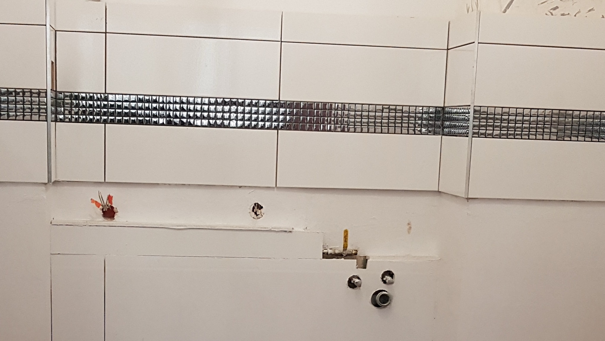 Ściana w trakcie układania płytek z dekoracyjnym pasem mozaiki, widoczne wystające kable elektryczne i rury hydrauliczne przed podłączeniem armatury.