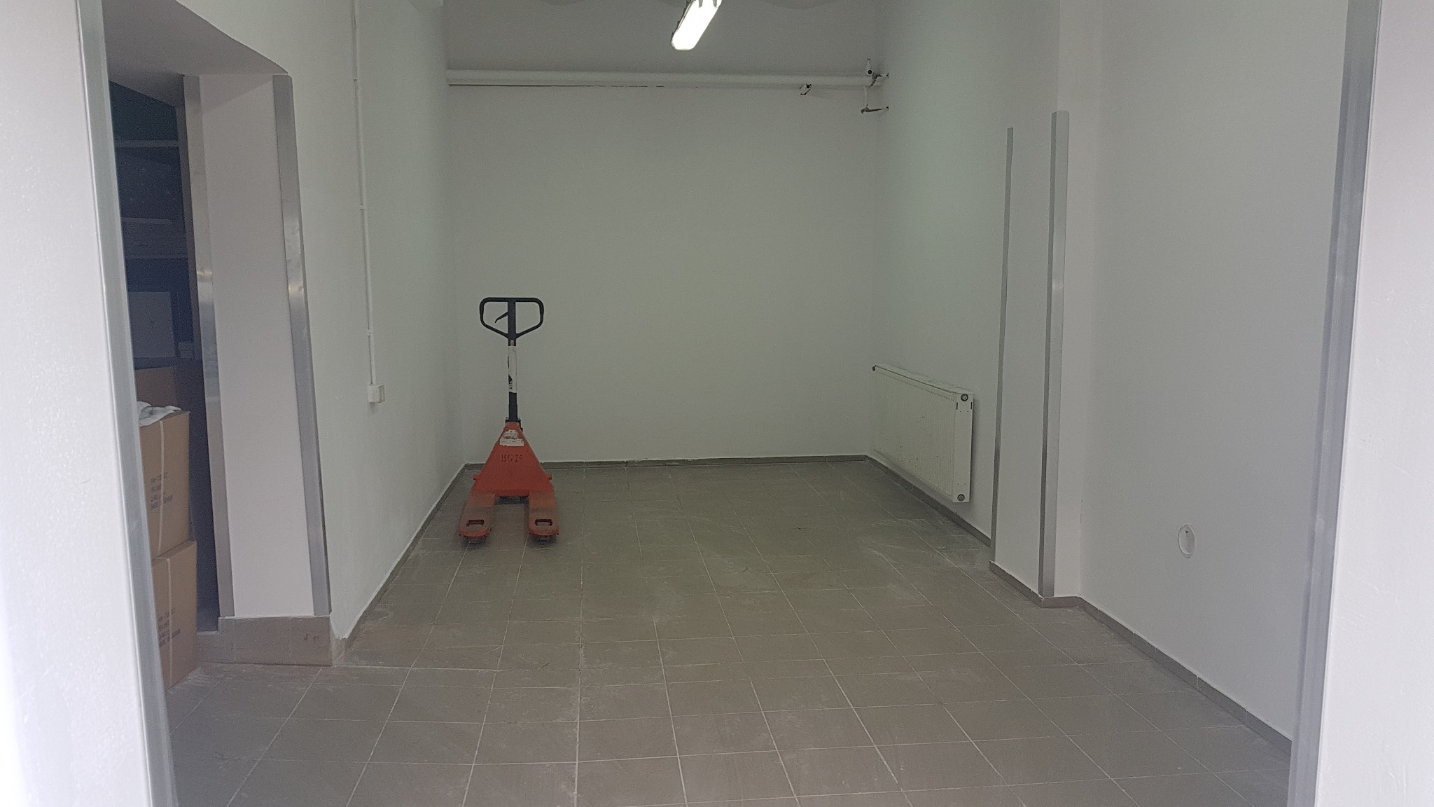 Puste pomieszczenie w trakcie prac wykończeniowych w Łodzi, z szarą, kwadratową podłogą z płytek, białymi ścianami, grzejnikiem i paleciakiem w kolorze pomarańczowym.