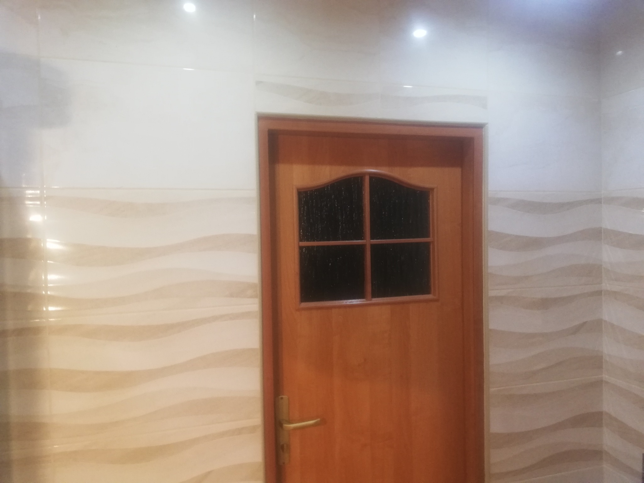 Drewniane drzwi z szybą w łazience wyłożonej płytkami z delikatnym wzorem fal, oświetlone punktowo.