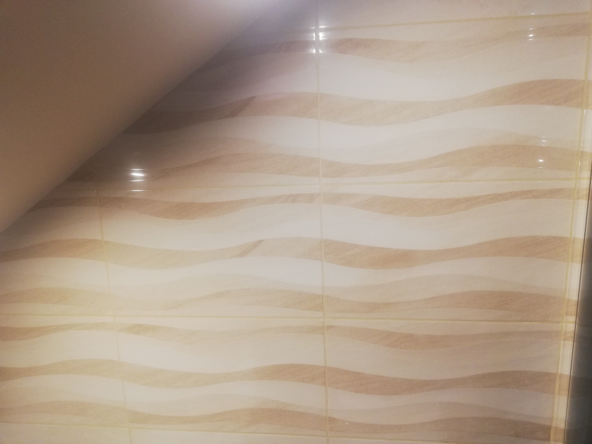 Ściana pokryta beżowo-białymi płytkami ceramicznymi z falistym wzorem, widoczne fugi, oświetlenie górne.