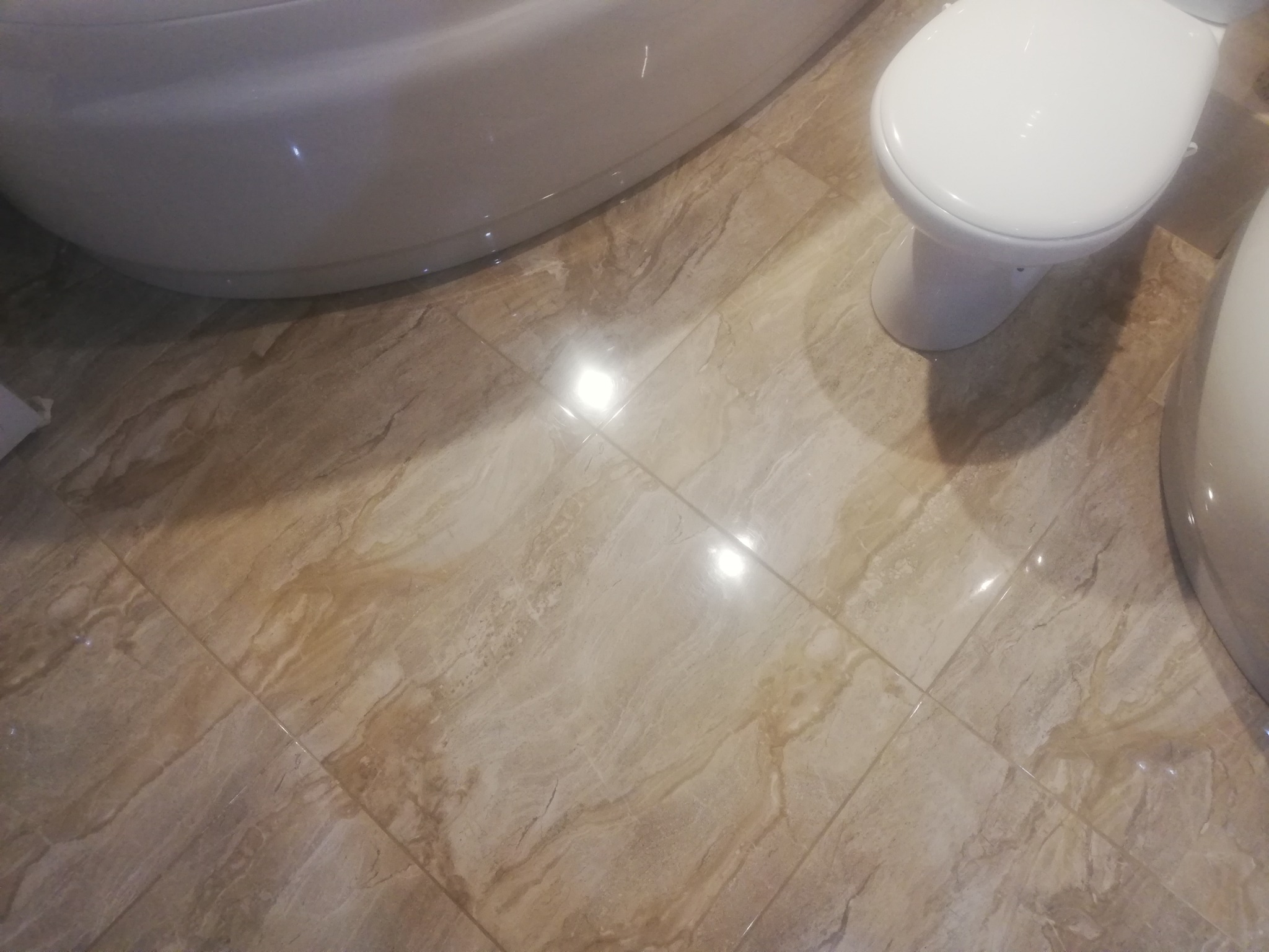 Beżowe, marmurkowe płytki podłogowe w łazience, widoczne fragmenty wanny i toalety, połyskujące od oświetlenia.