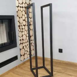 Metalowy, czarny stojak na drewno kominkowe o minimalistycznej, kubicznej formie, ustawiony na drewnianej podłodze w rogu pokoju, z dekoracyjną ścianą z drewna w tle.