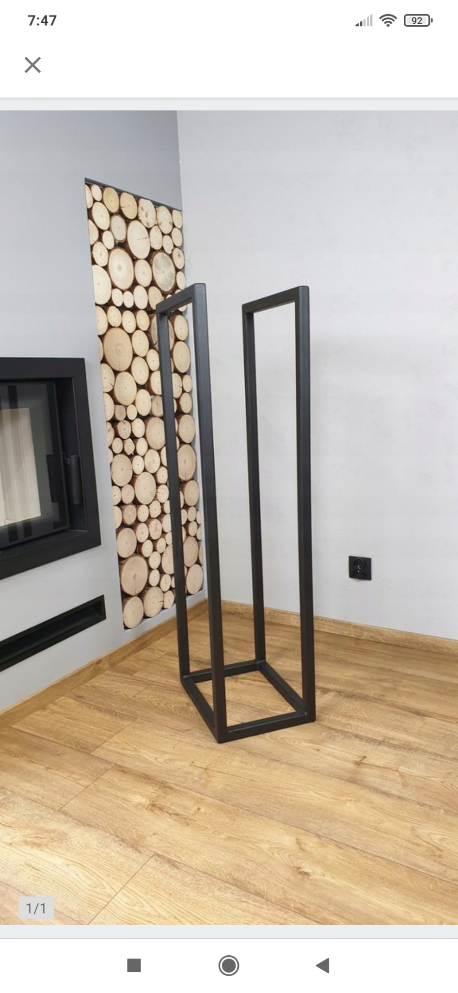 Metalowy, czarny stojak na drewno kominkowe o minimalistycznej, kubicznej formie, ustawiony na drewnianej podłodze w rogu pokoju, z dekoracyjną ścianą z drewna w tle.