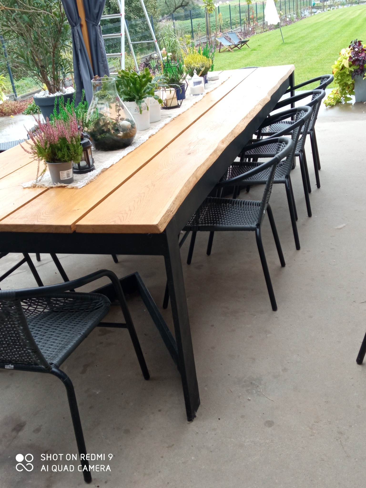 Długi, drewniany stół z czarnymi metalowymi nogami i krzesłami z plecionki na zewnątrz, dekorowany roślinami w doniczkach i szklanym naczyniu z roślinami, w tle trawnik i drzewa.