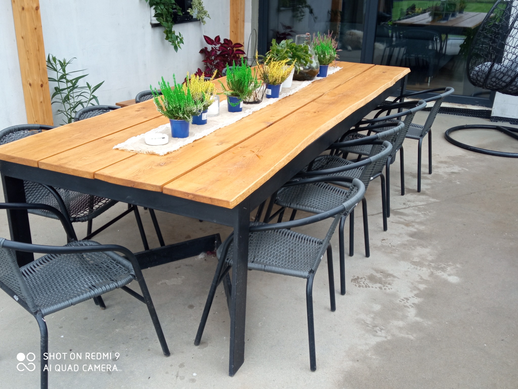 Długi drewniany stół z czarnymi metalowymi nogami i krzesłami z plecionego tworzywa sztucznego, ustawiony na zewnątrz i udekorowany rzędem doniczkowych wrzosów w kolorowych doniczkach.