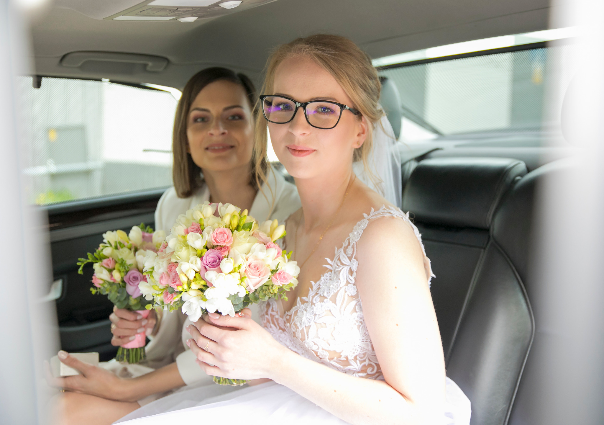 Panna młoda w okularach i druhna z bukietami kwiatów w samochodzie, widoczne skórzane siedzenia i zasłonięte okno.