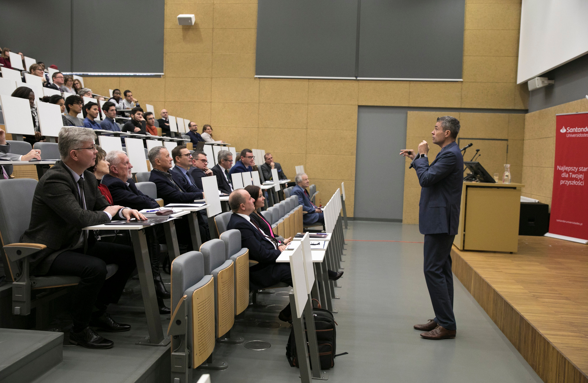 Prelegent w ciemnym garniturze gestykuluje, przemawiając do publiczności w auli podczas Dnia Internacjonalizacji na UMCS; widoczny baner Santander.