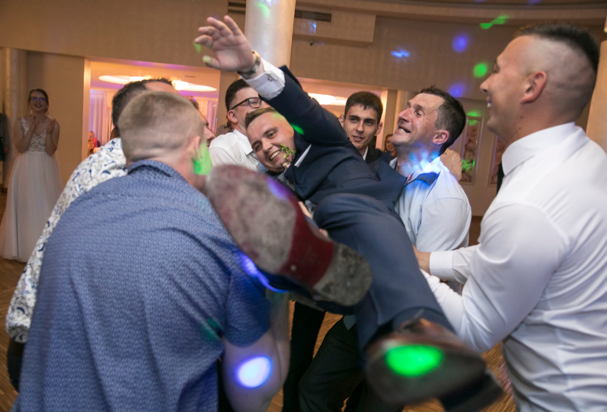 Pan młody niesiony na rękach przez gości weselnych podczas zabawy, w tle uśmiechnięta panna młoda w białej sukni.