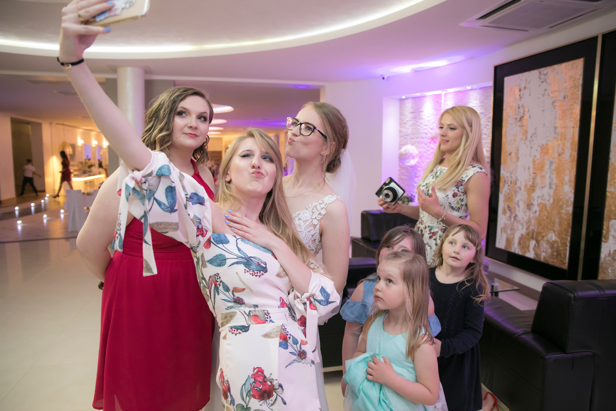 Grupa kobiet i dzieci pozuje do selfie w eleganckim wnętrzu, prawdopodobnie podczas uroczystości ślubnej, z panną młodą w tle.