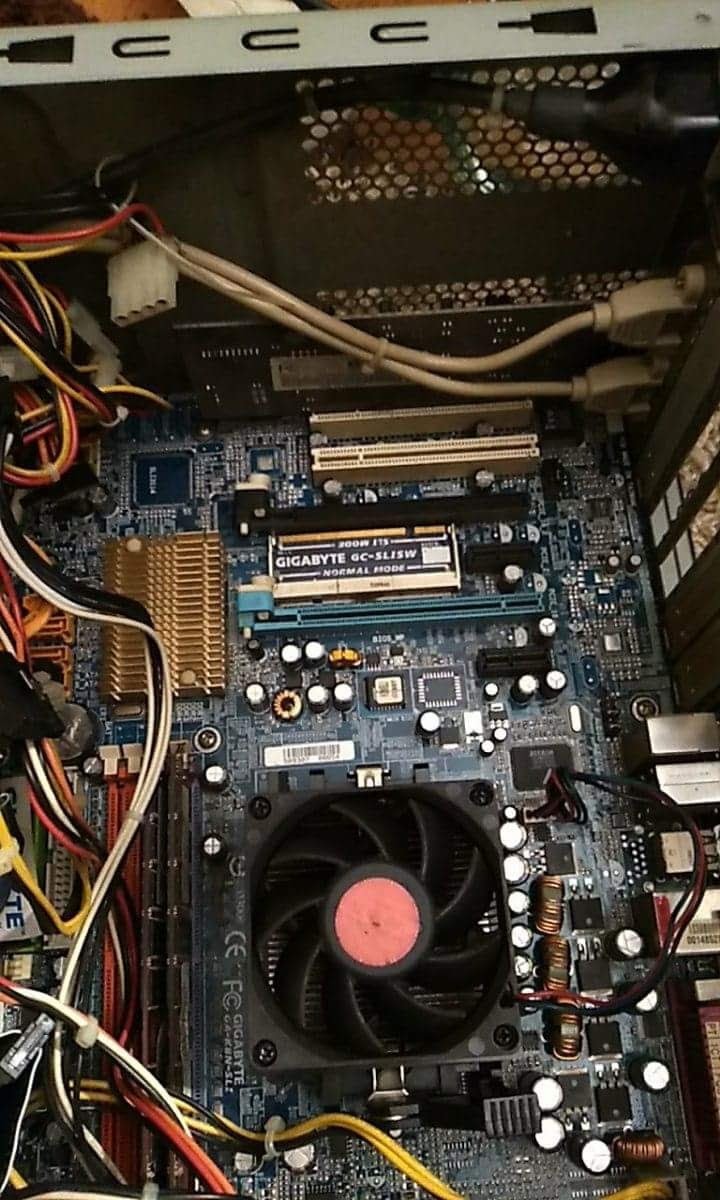 Otwarta obudowa komputera stacjonarnego, widoczna płyta główna Gigabyte z zamontowanym radiatorem procesora z wentylatorem z czerwoną naklejką na środku, liczne kable i złącza, w tle metalowa...