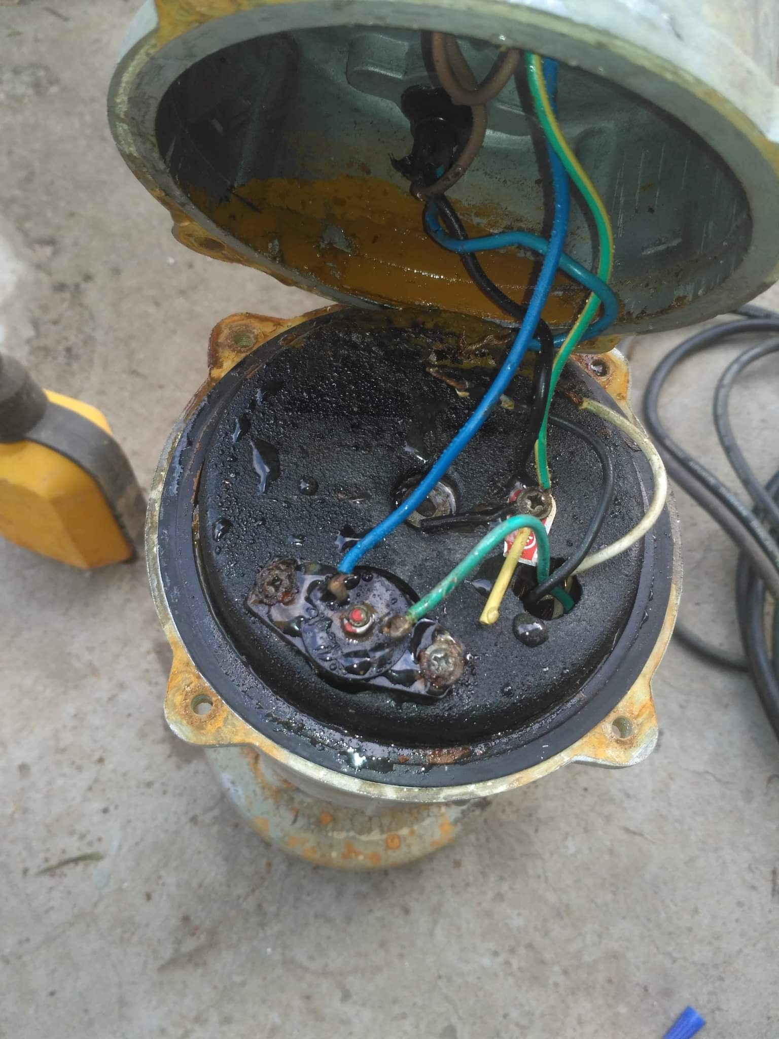 Otwarta, skorodowana puszka elektryczna z widocznymi, podłączonymi przewodami w kolorach: niebieskim, zielono-żółtym, białym i czarnym, zalana oleistą substancją.